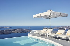 A poolside area with lounge chairs and an umbrella, overlooking a scenic coastline. - Katikies Chromata Santorini - The Leading Hotels of the World (Santorini)