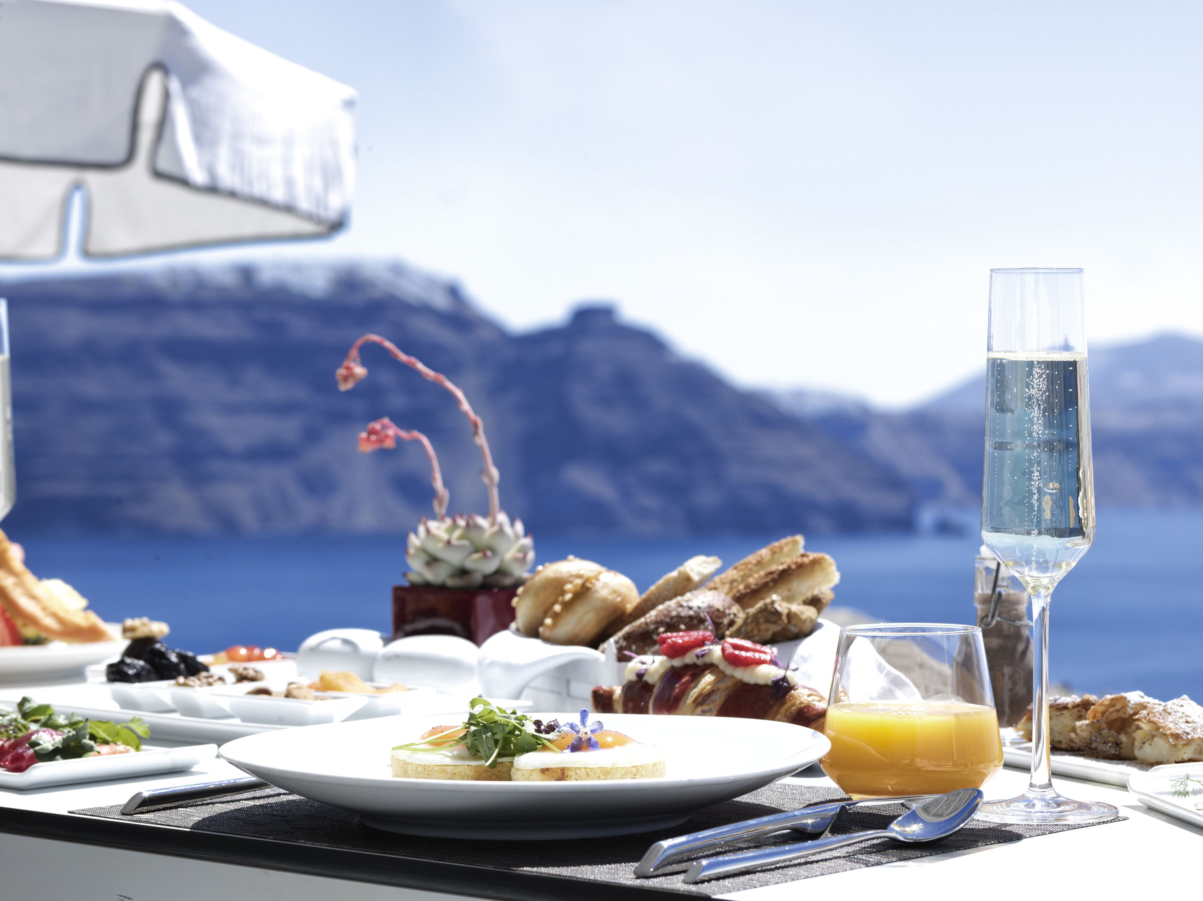 A table set with breakfast items including croissants, a glass of orange juice, and a glass of water, with a scenic mountain view in the background.