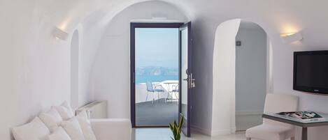 A modern, minimalist living room with a white sofa, a coffee table with a plant, a TV on a stand, and a view of the sea through an open door.