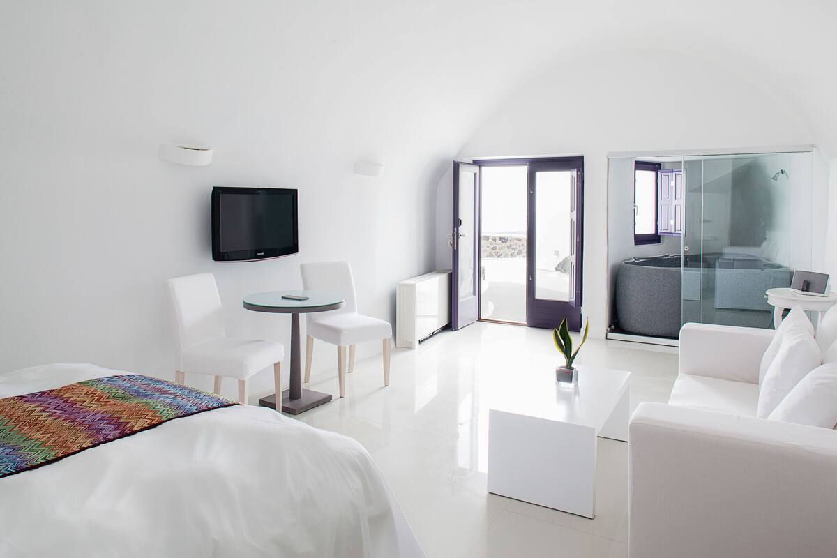 Ein modernes Hotelzimmer mit einem Bett, einem Fernseher, einem Balkon mit Glastür und einer Sitzecke mit Tisch und Stühlen.