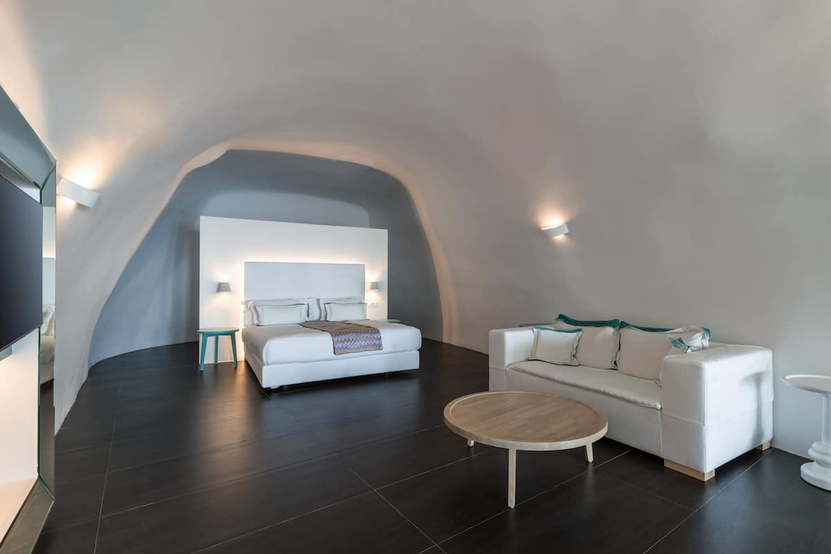 Ein modernes, minimalistisches Zimmer mit gewölbter Decke, einem weißen Sofa, einem runden Couchtisch aus Holz und einem Bett mit weißem Kopfteil.