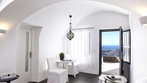 A modern, white-walled room with a curved ceiling, a dining area with a table and chairs, a small round table with a cup and book, and a view of the sea through an open door. - Katikies Chromata Santorini - The Leading Hotels of the World (Santorini)