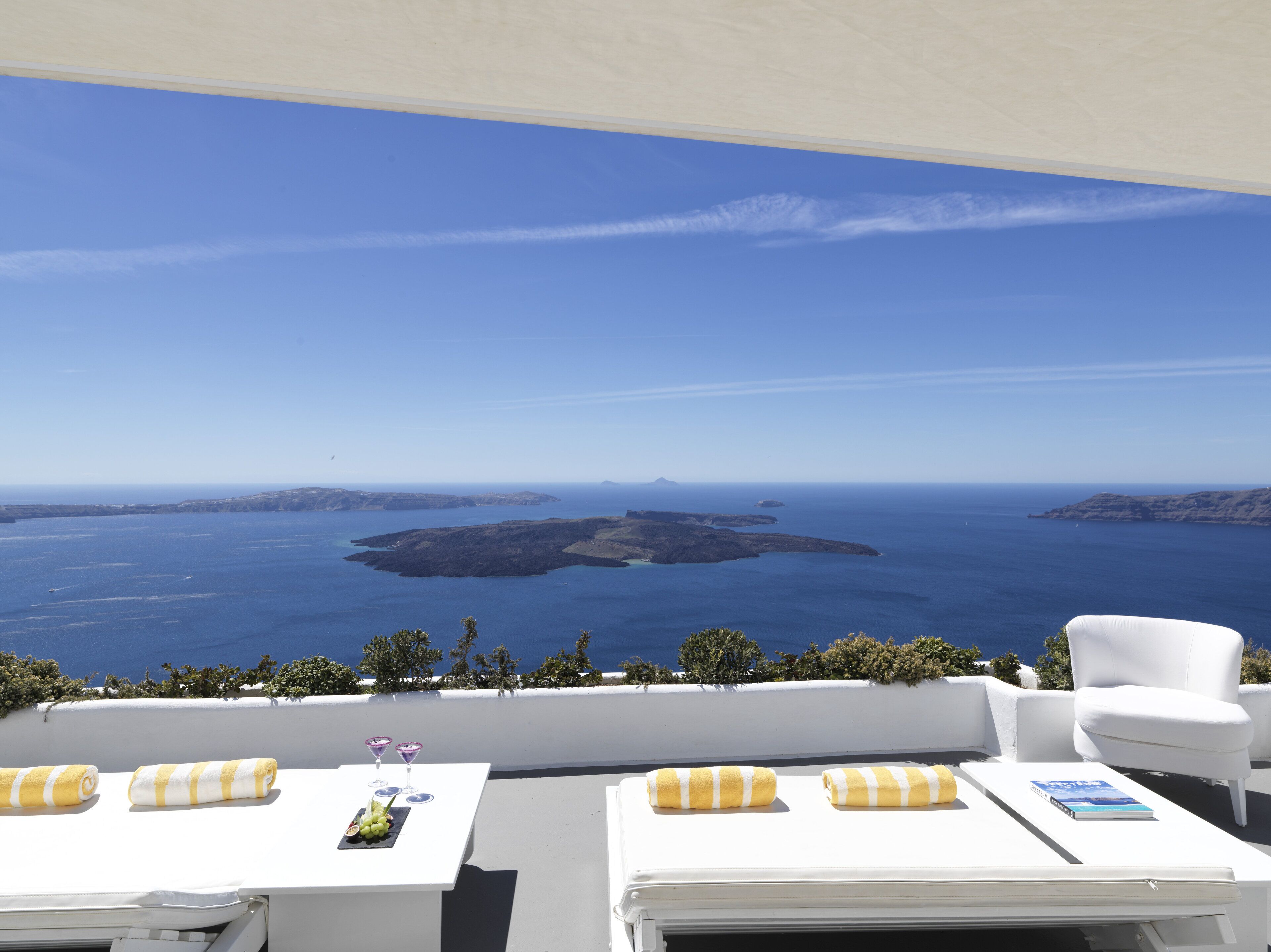 Une terrasse avec des meubles blancs, une table sur laquelle se trouvent un verre à martini et un livre, et une vue sur la mer et les îles.