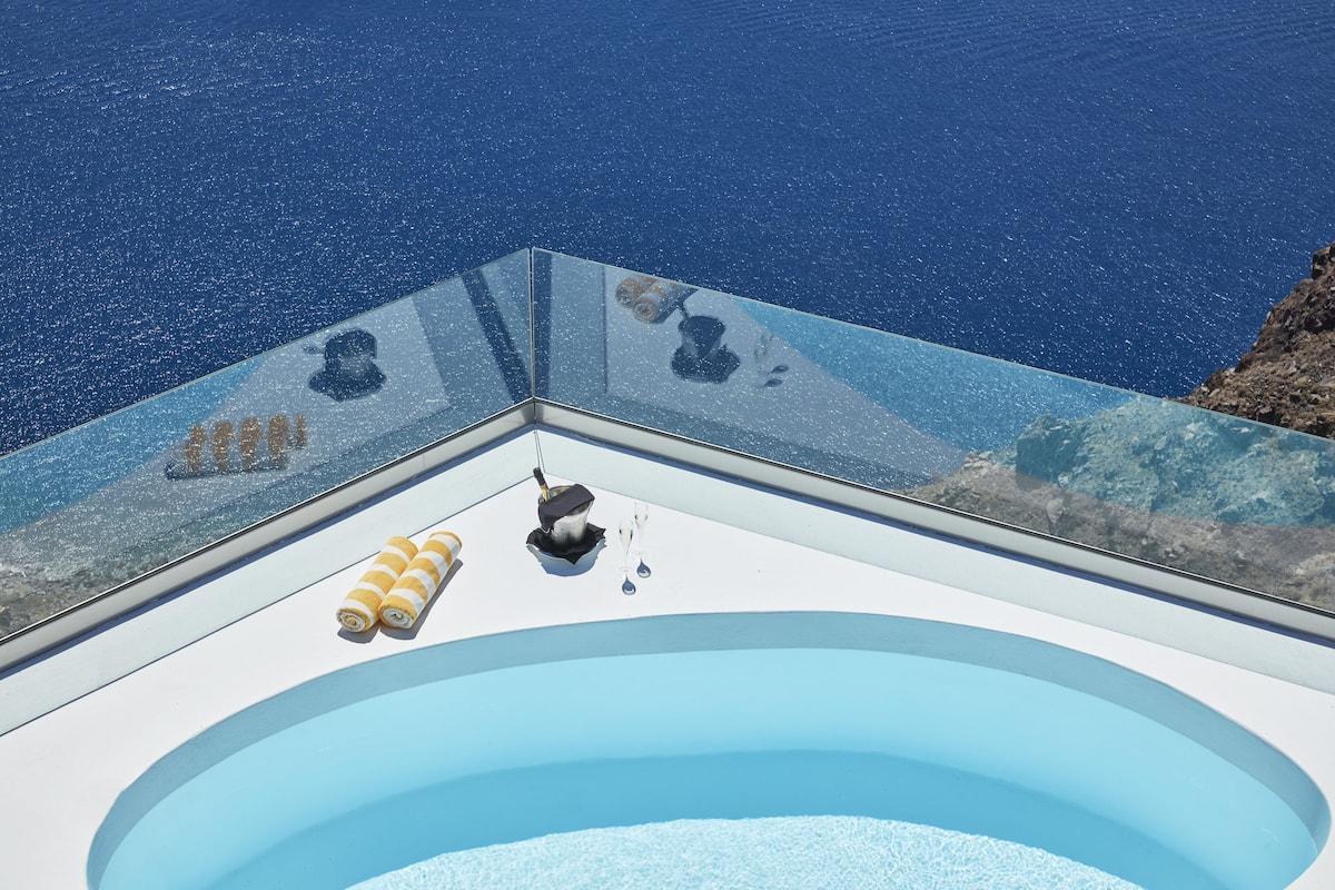 Ein glasüberdachter Infinity-Pool mit freiem Blick auf das Meer, eine kleine Sitzecke mit Kissen sowie eine Champagnerflasche.