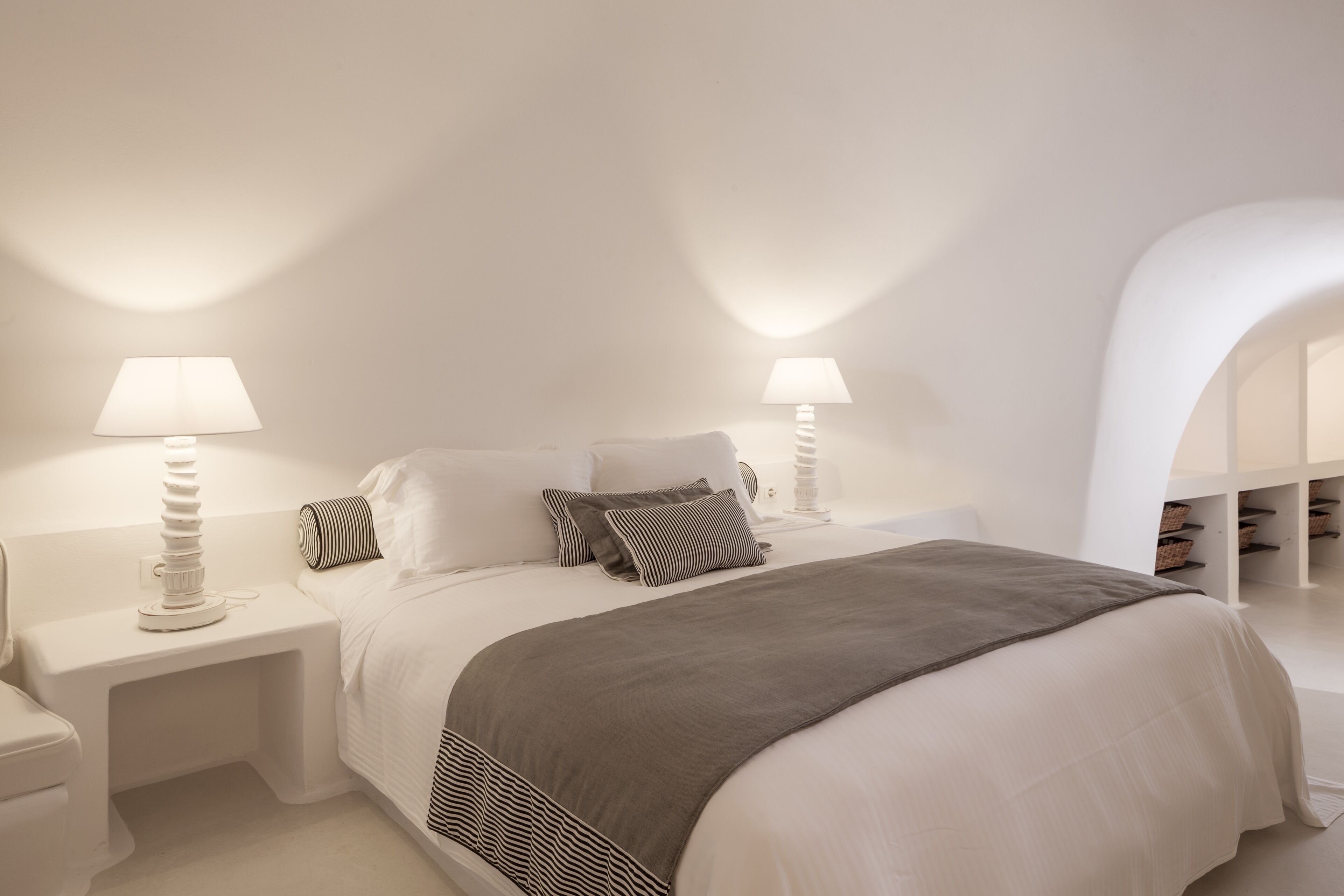 A modern bedroom with a large bed, two bedside tables with lamps, a grey throw blanket, and a white archway leading to a storage area.