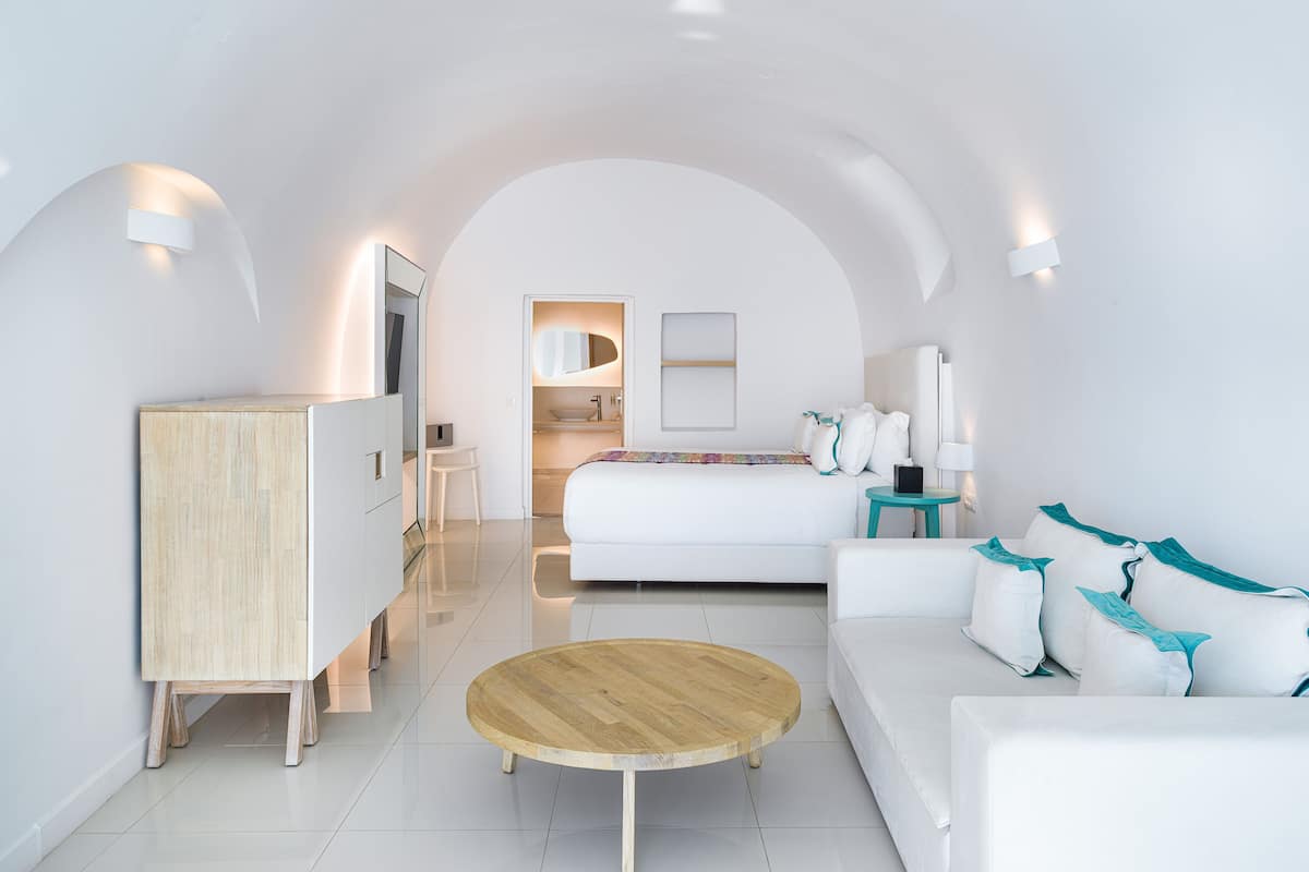 Ein modernes Schlafzimmer in Weiß mit einem Bett, einem Holztischchen, einem weißen Sofa und einem Schreibtisch.