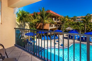 A pool area with blue umbrellas, lounge chairs, and a building with balconies and palm trees in the background.