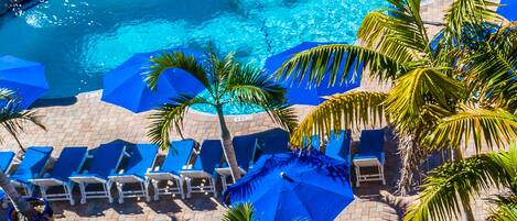 A pool area with blue umbrellas, lounge chairs, and palm trees.