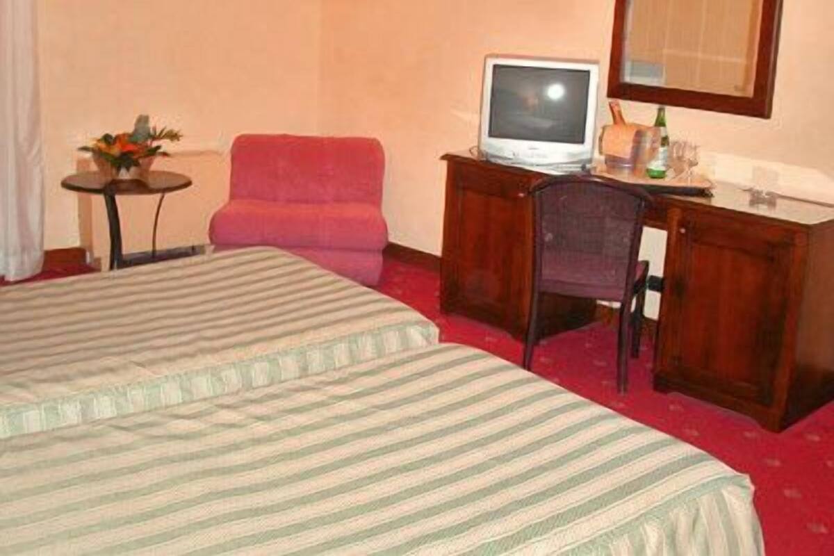 A hotel room with a bed, a desk with a computer, a chair, and a small table with a vase of flowers.