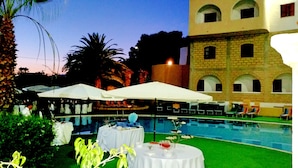 A poolside area with tables and umbrellas, a pool, and a building with arched windows. - Hotel Akrabello (Agrigento)