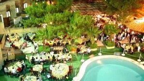 An outdoor dining area with multiple round tables and chairs, surrounded by a pool and palm trees. - Hotel Akrabello (Agrigento)