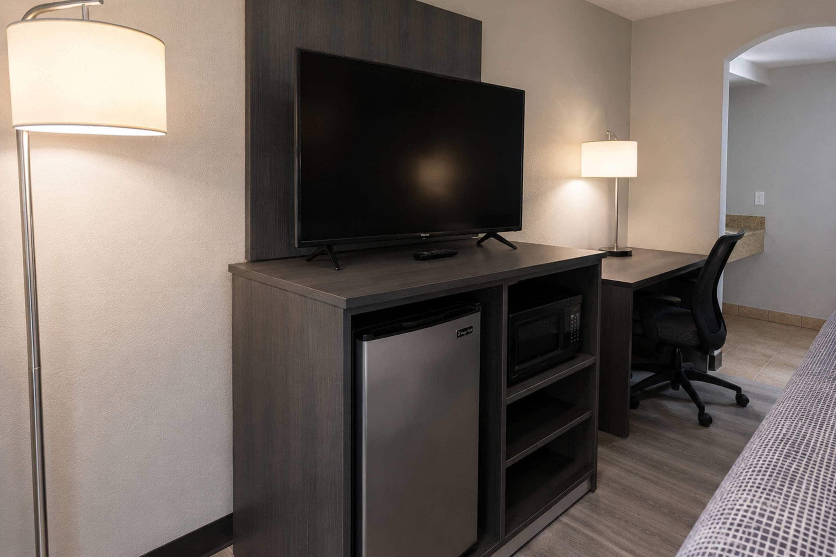 Habitación de hotel con televisión de pantalla plana sobre un soporte de madera, un mini refrigerador, un escritorio con silla y dos lámparas de noche.