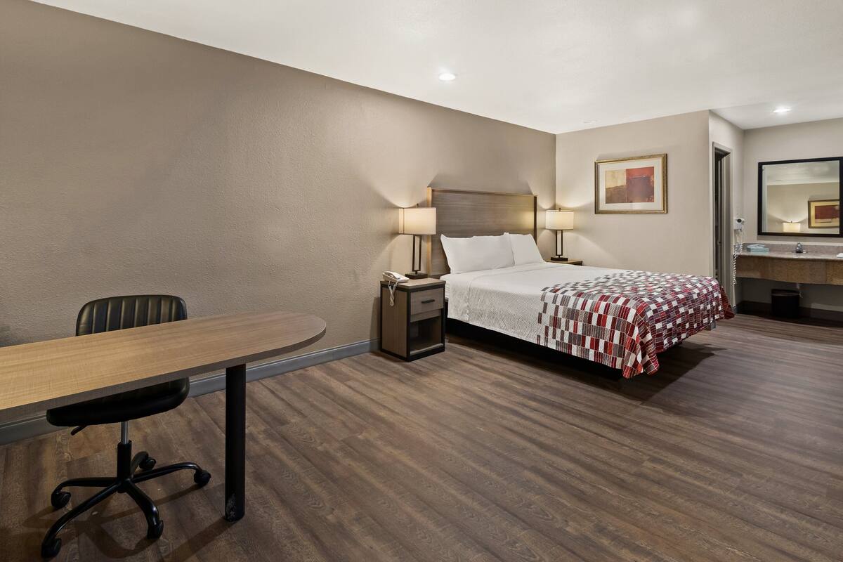 A hotel room with a bed, bedside table, lamp, chair, and a desk.