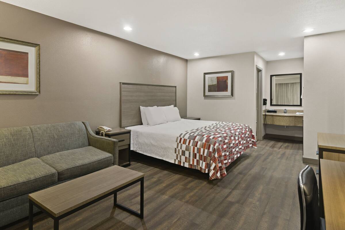 A hotel room with a bed, a sofa, a small table, a nightstand, a mirror, and a bathroom visible through an open door.