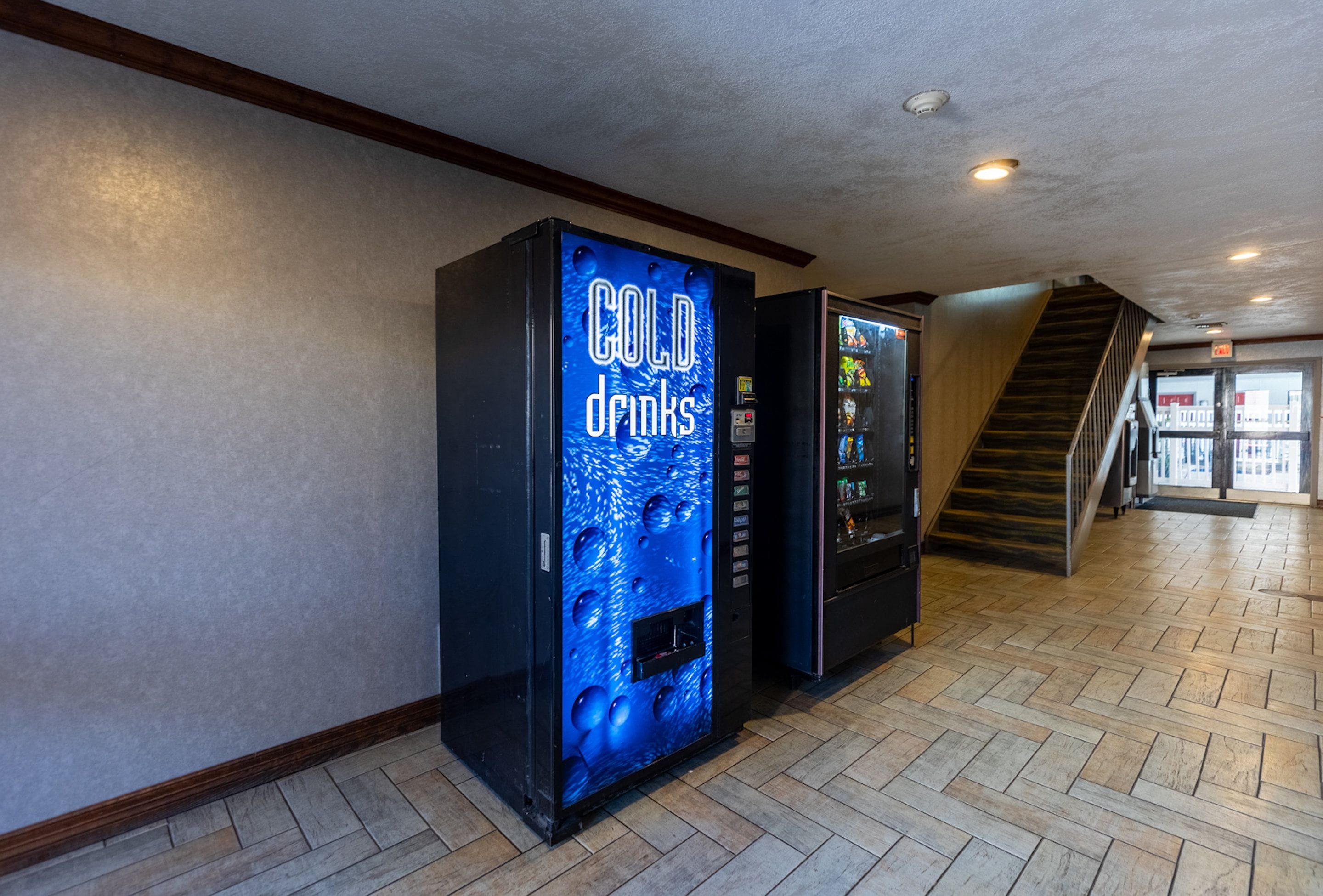 A hallway with two vending machines, one labeled 'COLD drinks' and the other with a colorful display. A wooden staircase is visible in the background.