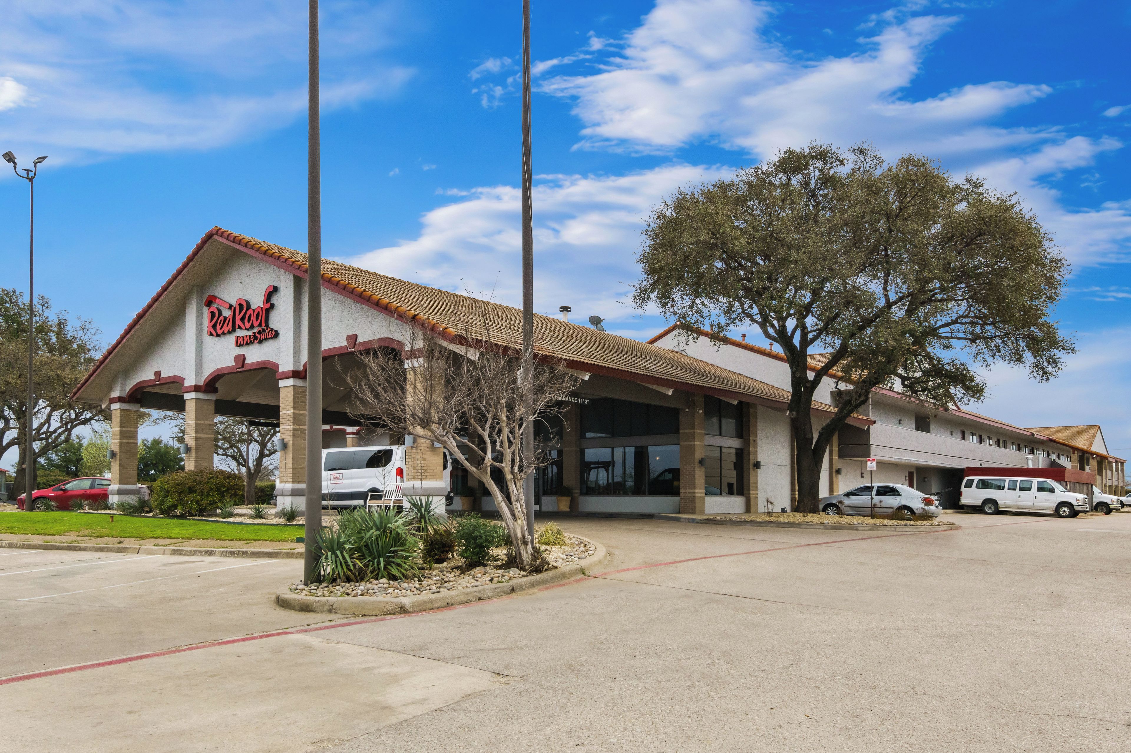 A Red Roof Inn & Suites with a tiled roof, parking area, and a large tree in front.