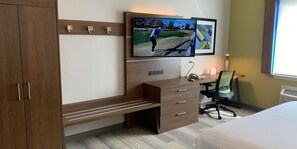 A hotel room with a large flat-screen TV mounted on the wall, a wooden desk with a chair, a bench, and a bed. - Holiday Inn Express & Suites Atlanta - Tucker Northlake by IHG (Tucker)
