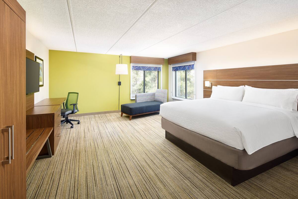 A hotel room with a large bed, a desk, a chair, a bench, and a window with curtains.
