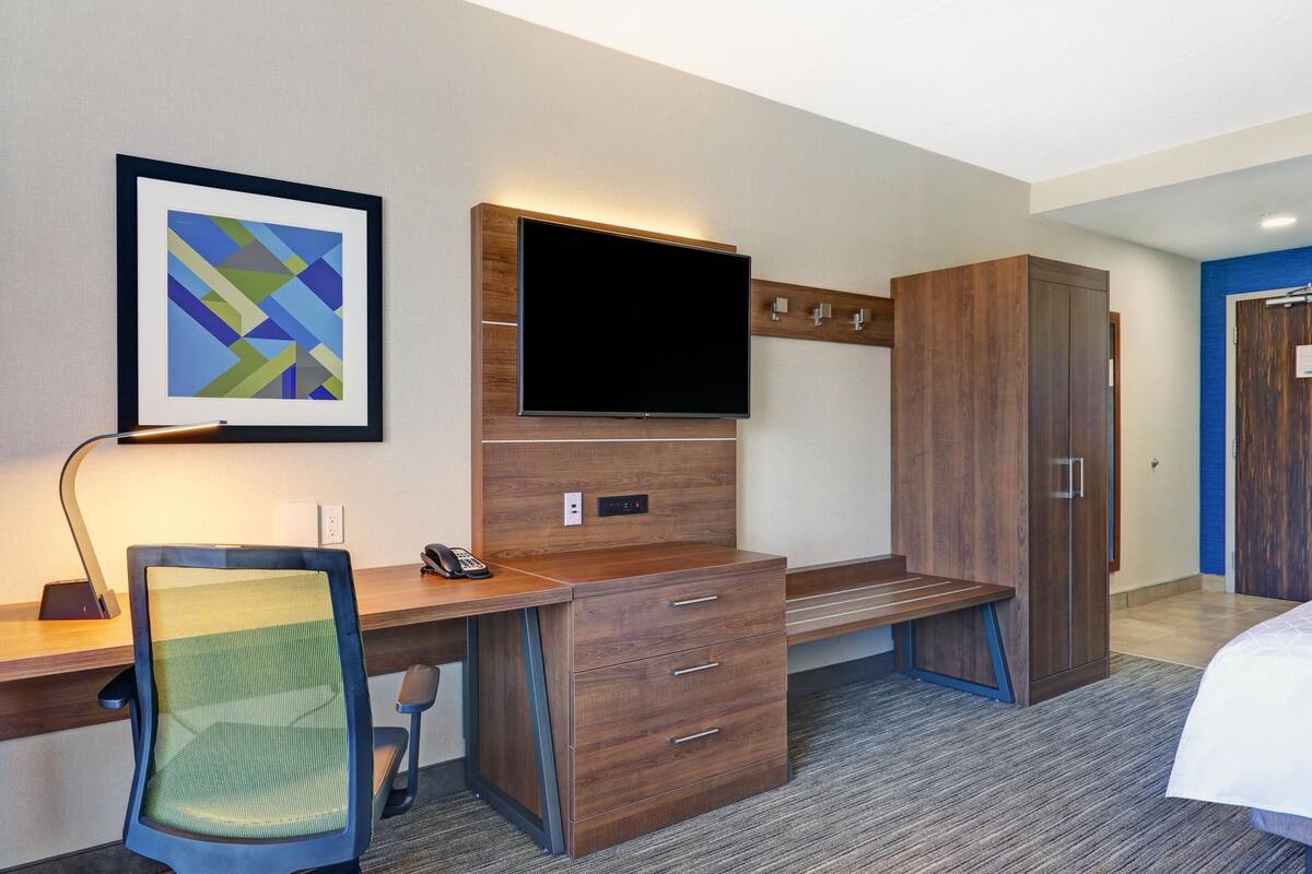 A hotel room with a wooden desk, a chair, a flat-screen TV, a wardrobe, and a bed.