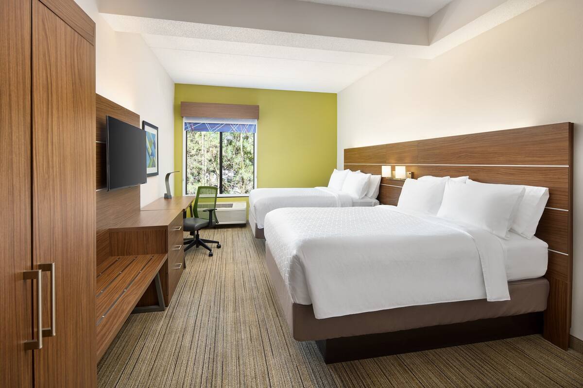 A hotel room with two beds, a desk with a flat-screen TV, and a view of greenery outside.