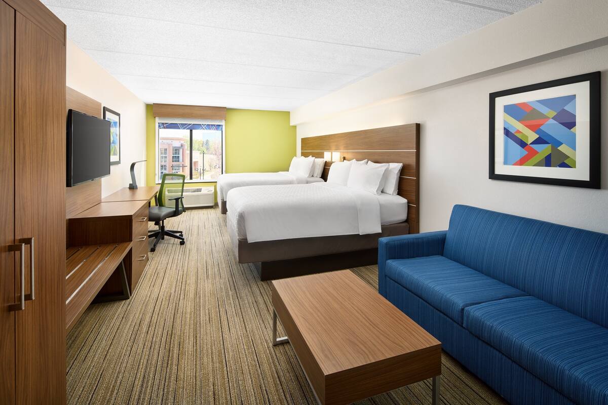 A hotel room with two beds, a desk, a blue sofa, a TV, and a window with a view of buildings.