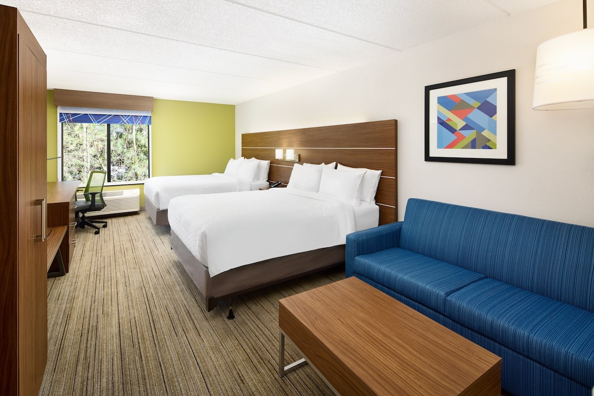 A hotel room with two beds, a blue sofa, a wooden desk, and a window with a view of trees.