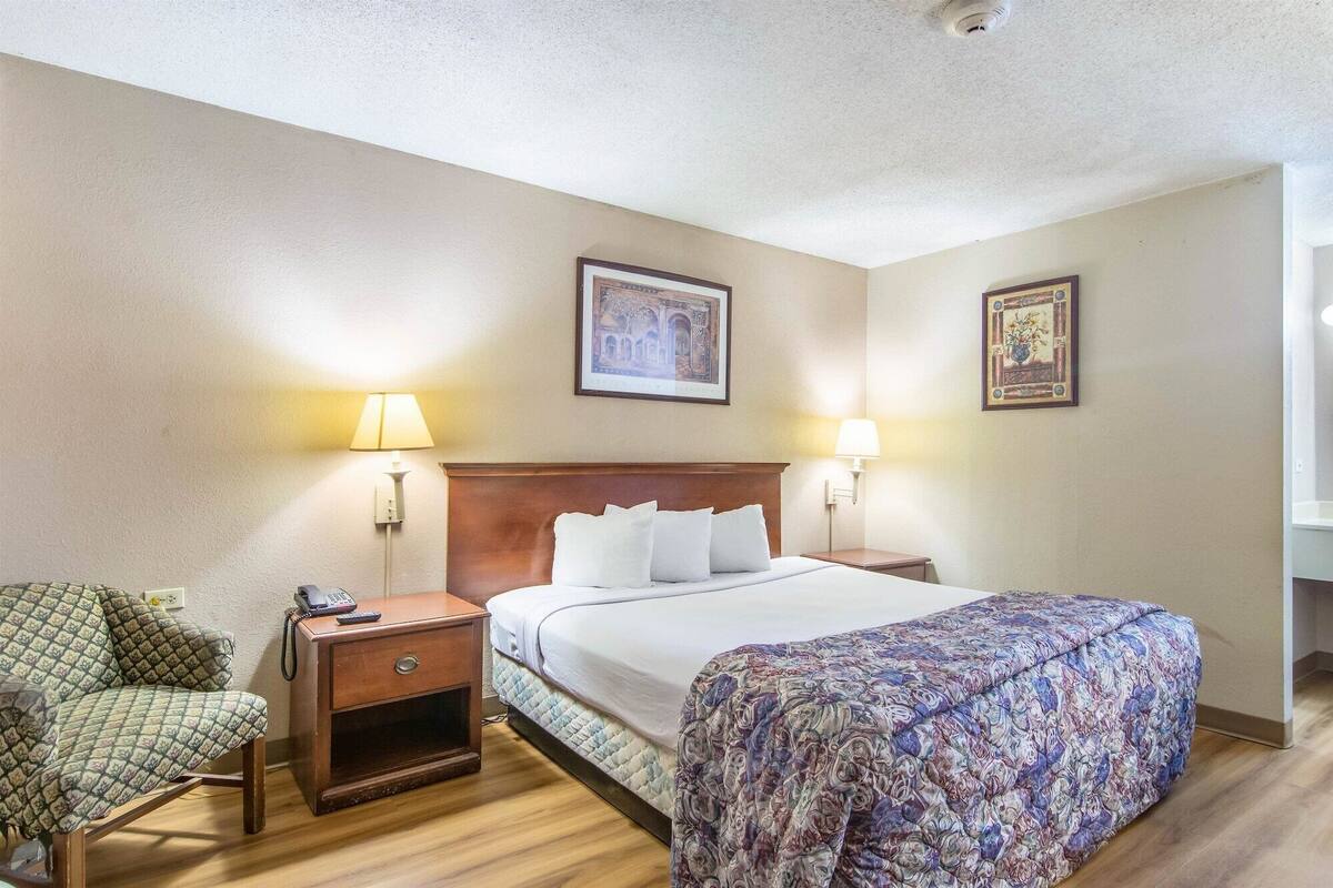 A hotel room with a bed, two bedside lamps, a nightstand with a phone, a chair, and two wall-mounted pictures.