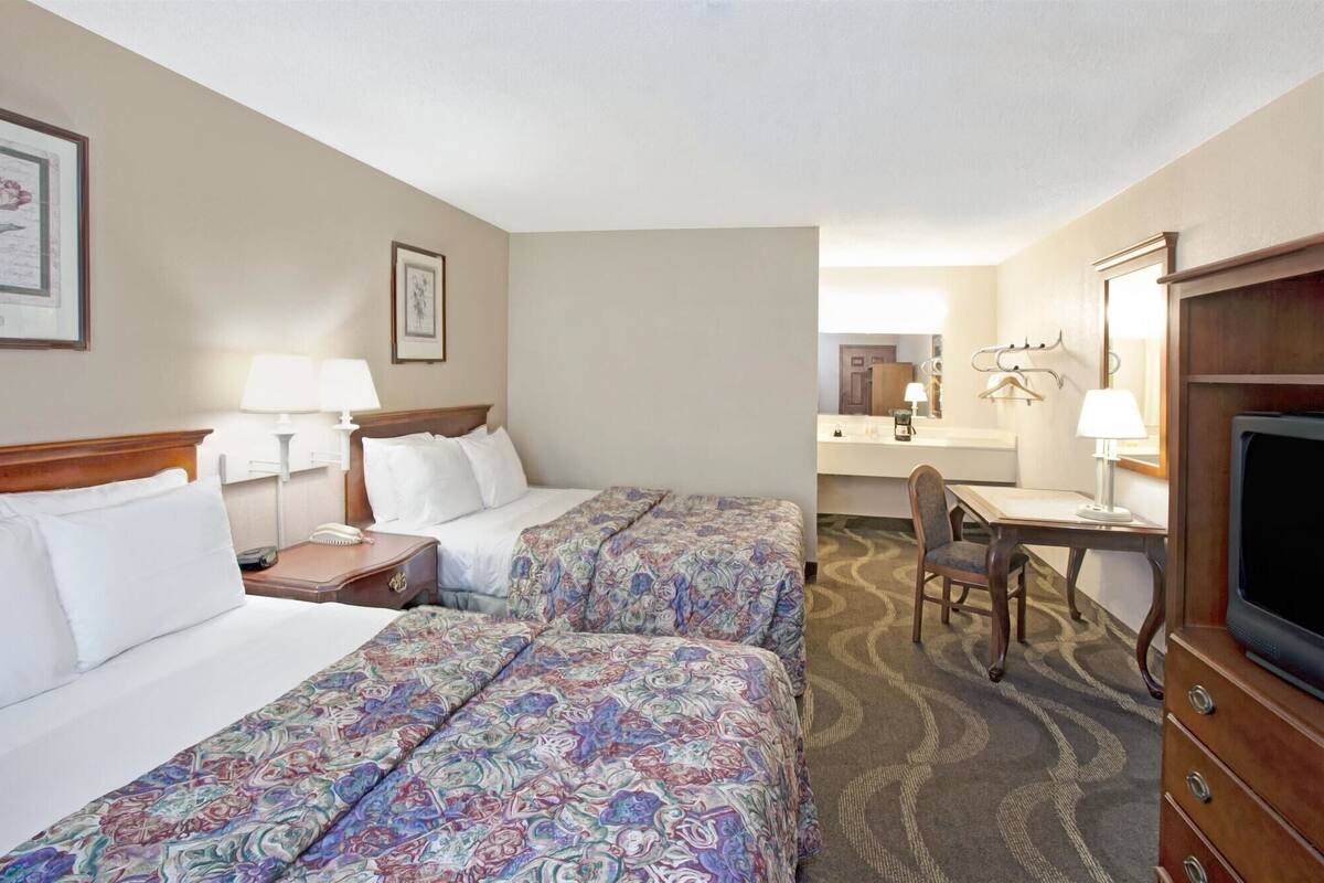 A hotel room with two beds, a desk, a chair, a television, and a bathroom visible in the background.