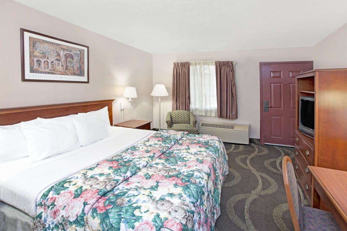 A hotel room with a large bed, a chair, a desk with a television, a window with curtains, and a framed picture on the wall.