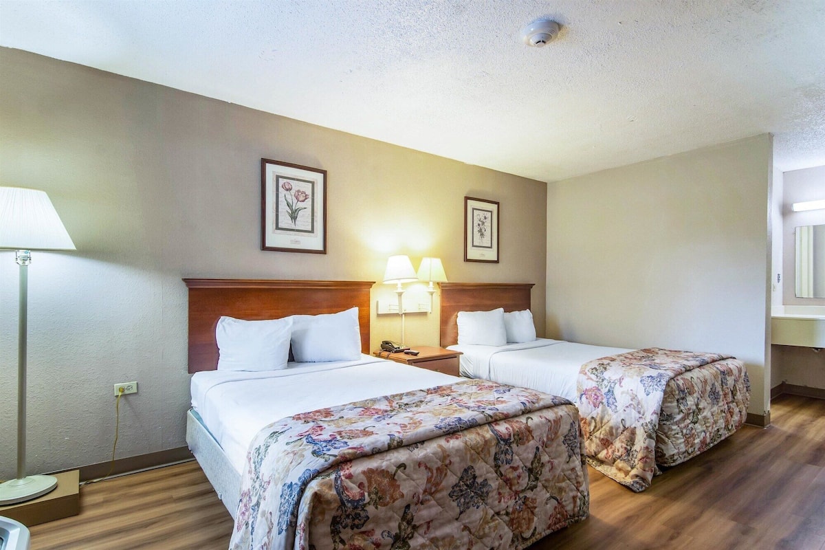 A hotel room with two beds, a nightstand with a lamp, and framed floral artwork on the walls.