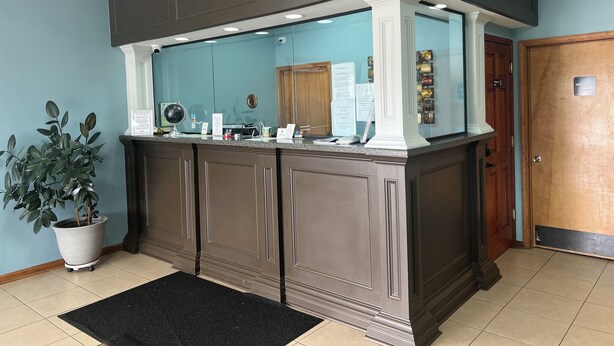 A reception desk with a glass partition, a potted plant, and a black doormat. - American Inn  - Pontotoc (Pontotoc)