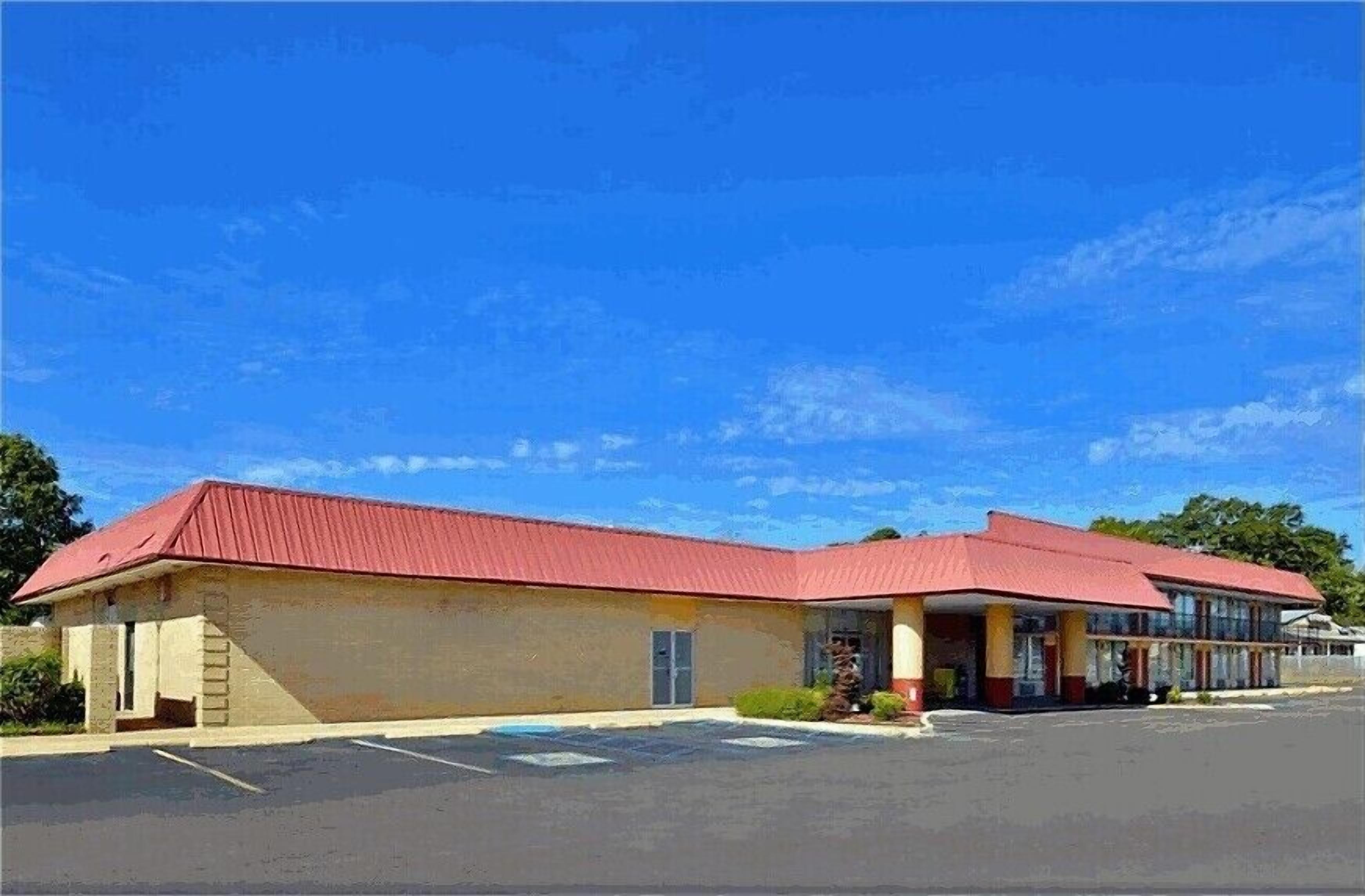A two-story motel with a red roof and beige walls, featuring a parking lot and a clear blue sky.