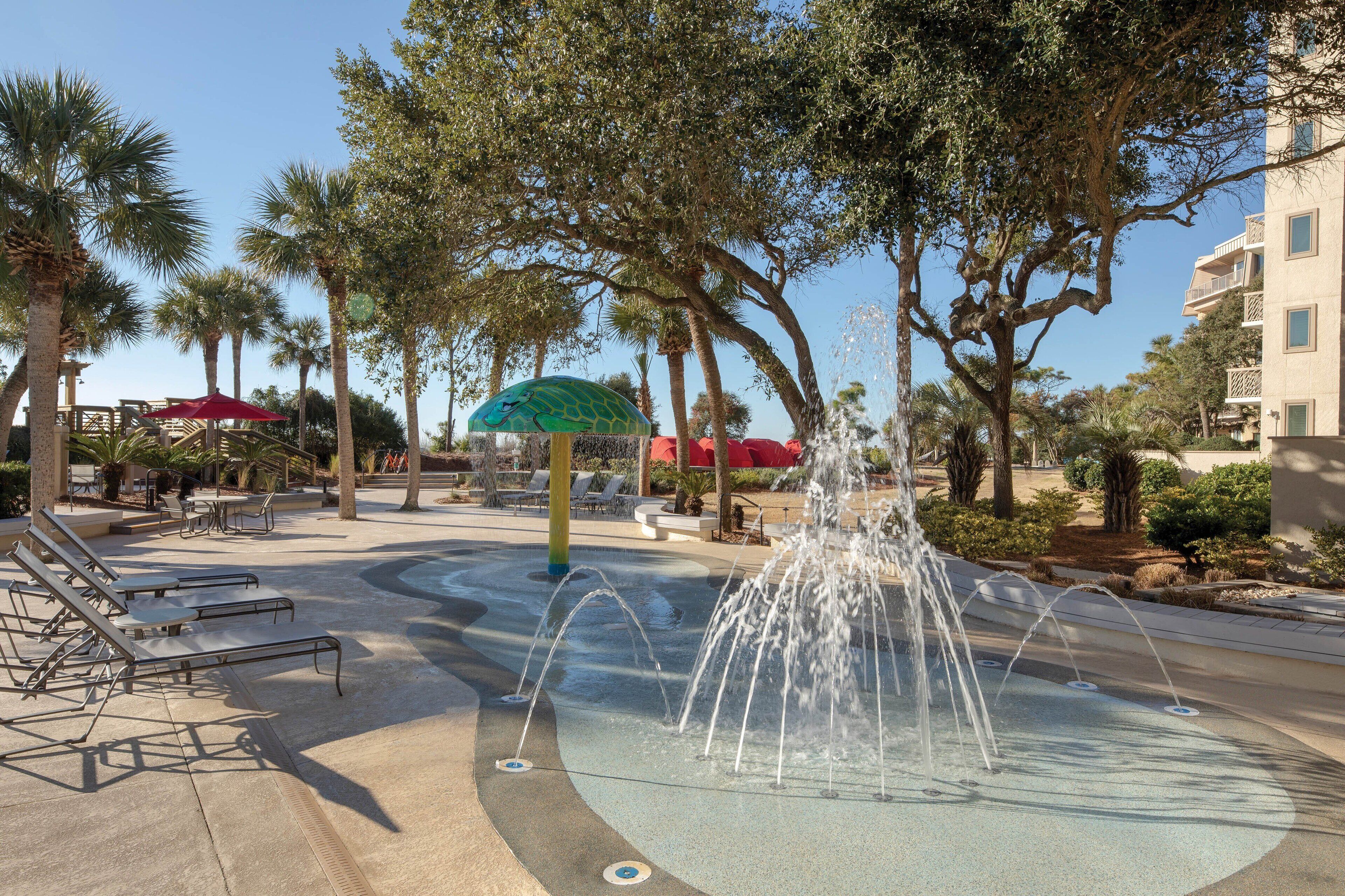 A water fountain surrounded by palm trees and lounge chairs in a courtyard.