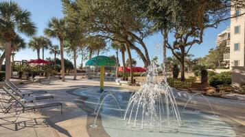 A water fountain surrounded by palm trees and lounge chairs in a courtyard.