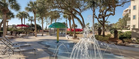 Une fontaine d’eau, entourée de palmiers et de chaises longues, se trouve dans une cour.