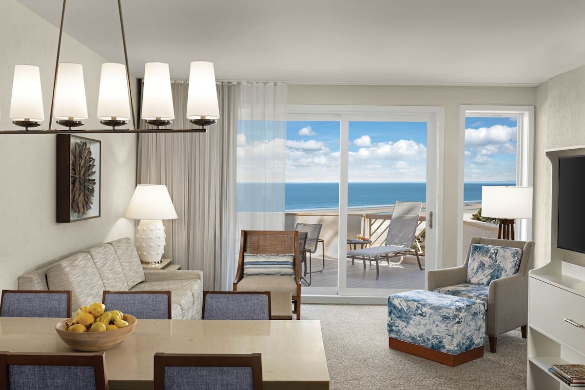 A hotel room with a balcony, a dining area, and a seating area with a view of the ocean.