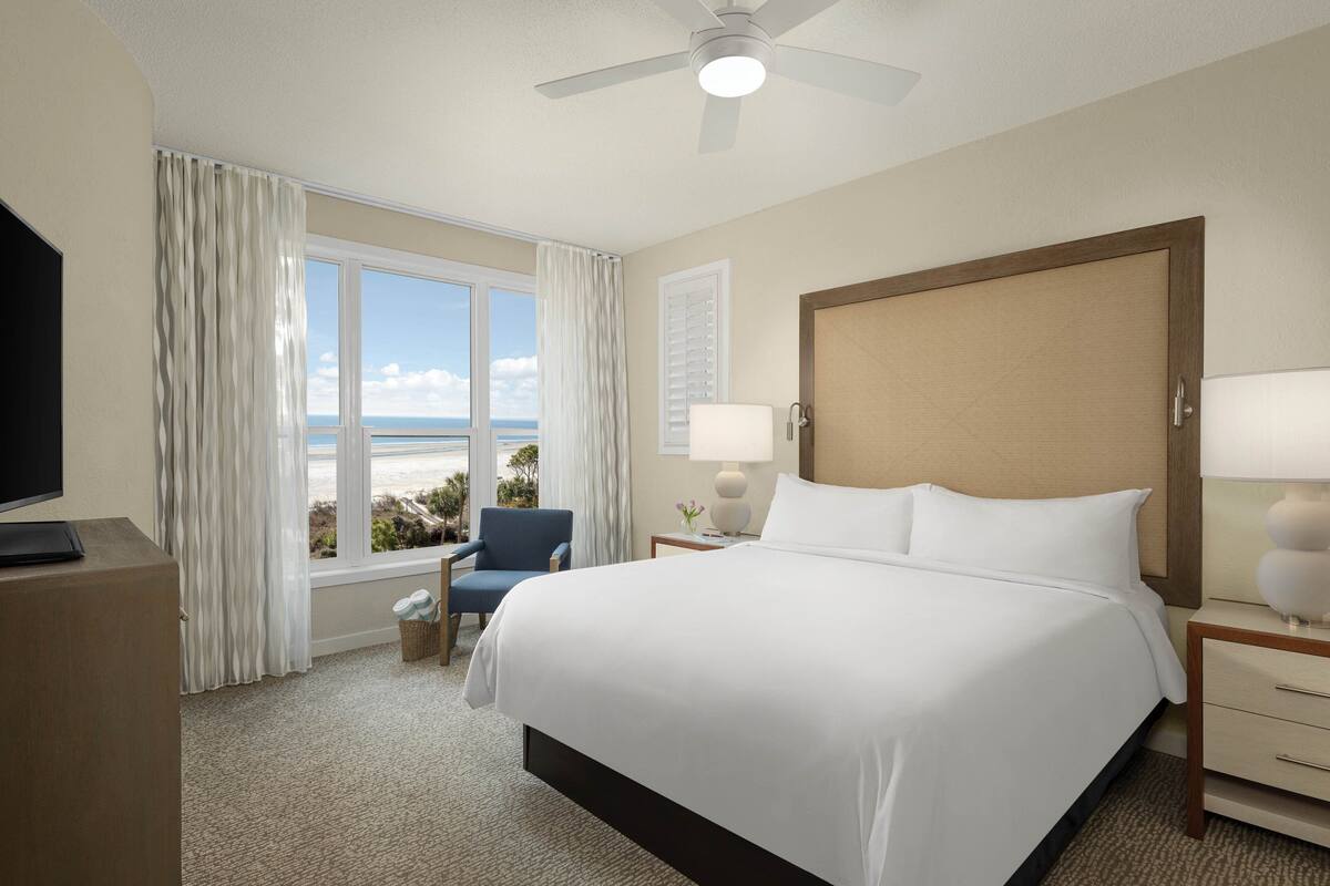 A hotel room with a large bed, a TV, a chair, a window with a view of the beach, and a ceiling fan.