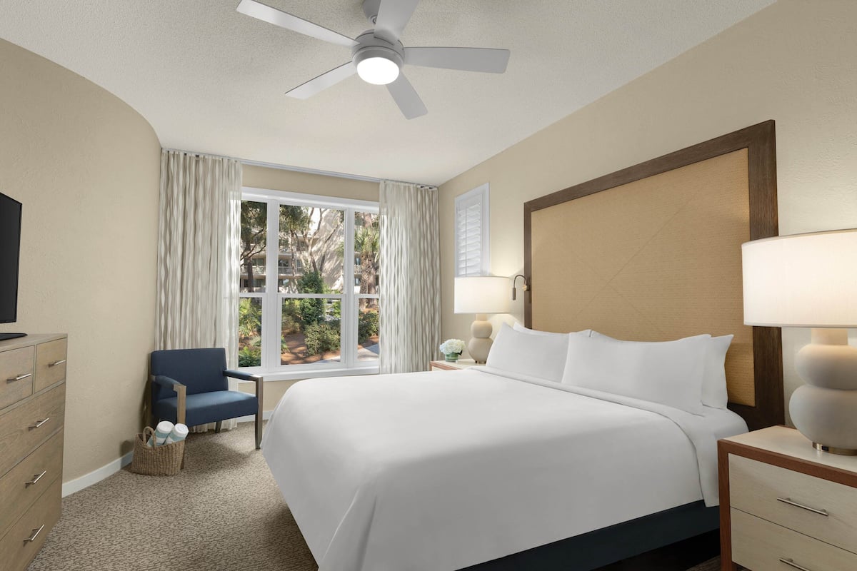 A hotel room with a large bed, a ceiling fan, a window with curtains, a desk with a lamp, and a TV on a dresser.
