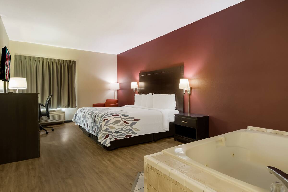 Habitación de hotel con cama, lámparas de noche, escritorio con silla, ventana grande con cortinas y bañera.