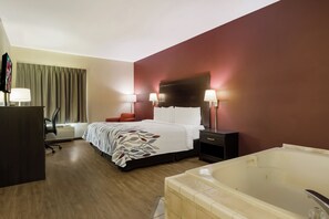 Ein Hotelzimmer mit Bett, Schreibtisch, Stuhl, Schreibtischlampe, Nachttisch mit Lampe, einem roten Sessel, einem großen Fenster mit Vorhängen und einer Badewanne.