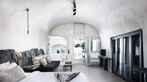A modern hotel room with a large bed, a sofa, and a desk. There is a view of the outside through an open door. - On The Rocks Hotel (Santorini)