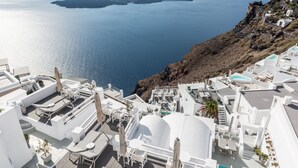 A coastal hotel with white architecture, outdoor seating areas, and a view of the sea and islands. - On The Rocks Hotel (Santorini)