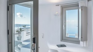 A modern hotel room with a glass door leading to a balcony, a bedside table with a tablet, and a view of the sea. - On The Rocks Hotel (Santorini)