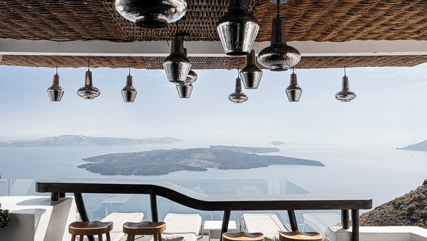 A terrace with a view of the sea, hanging pendant lights, and bar stools. - On The Rocks Hotel (Santorini)