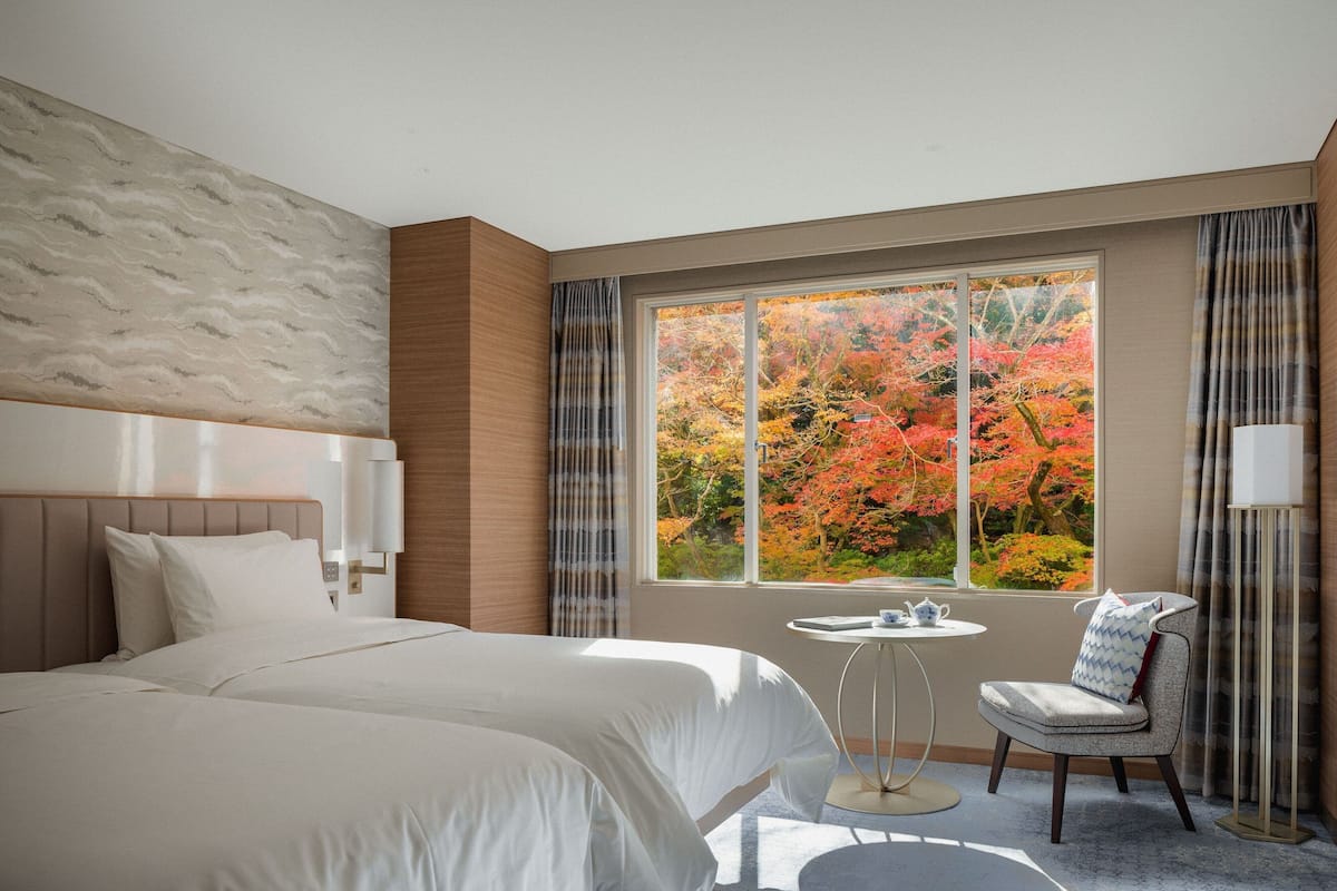 A hotel room with a large bed, a small round table, a chair, and a window with autumn foliage view.