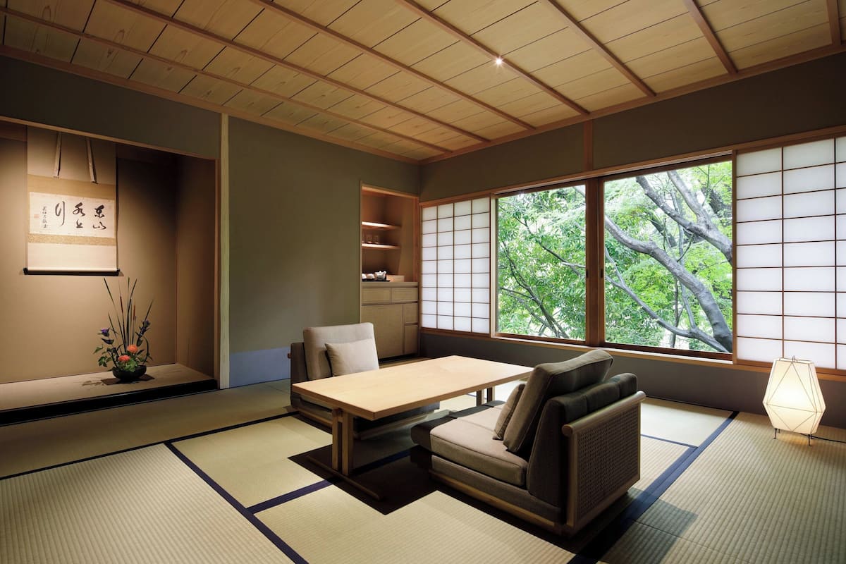 A traditional Japanese room with tatami flooring, a low table, a sofa, and a view of trees through large windows.