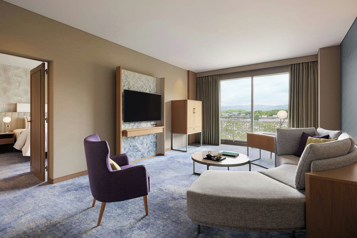 A hotel room with a large window, a flat-screen TV, a sofa, a chair, and a desk.