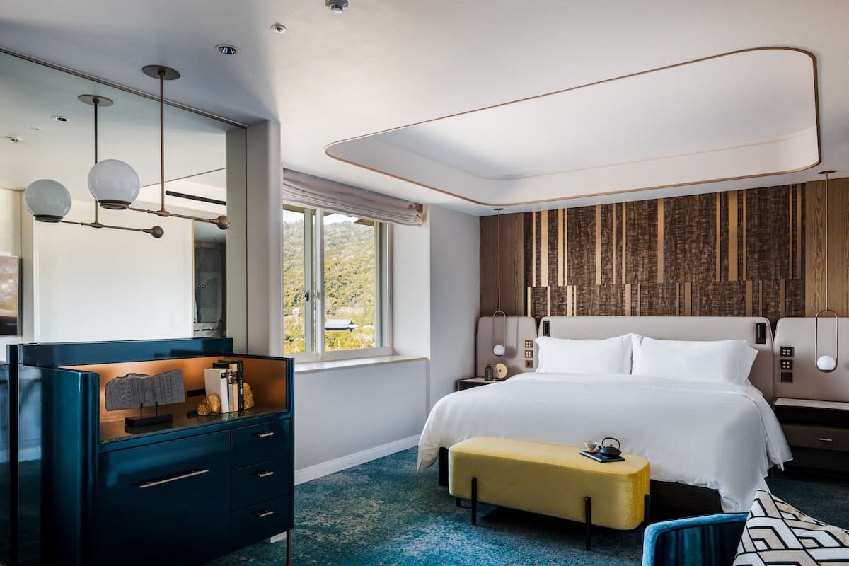 A modern hotel room with a large bed, a desk with drawers, a bench, and a view of the outdoors.