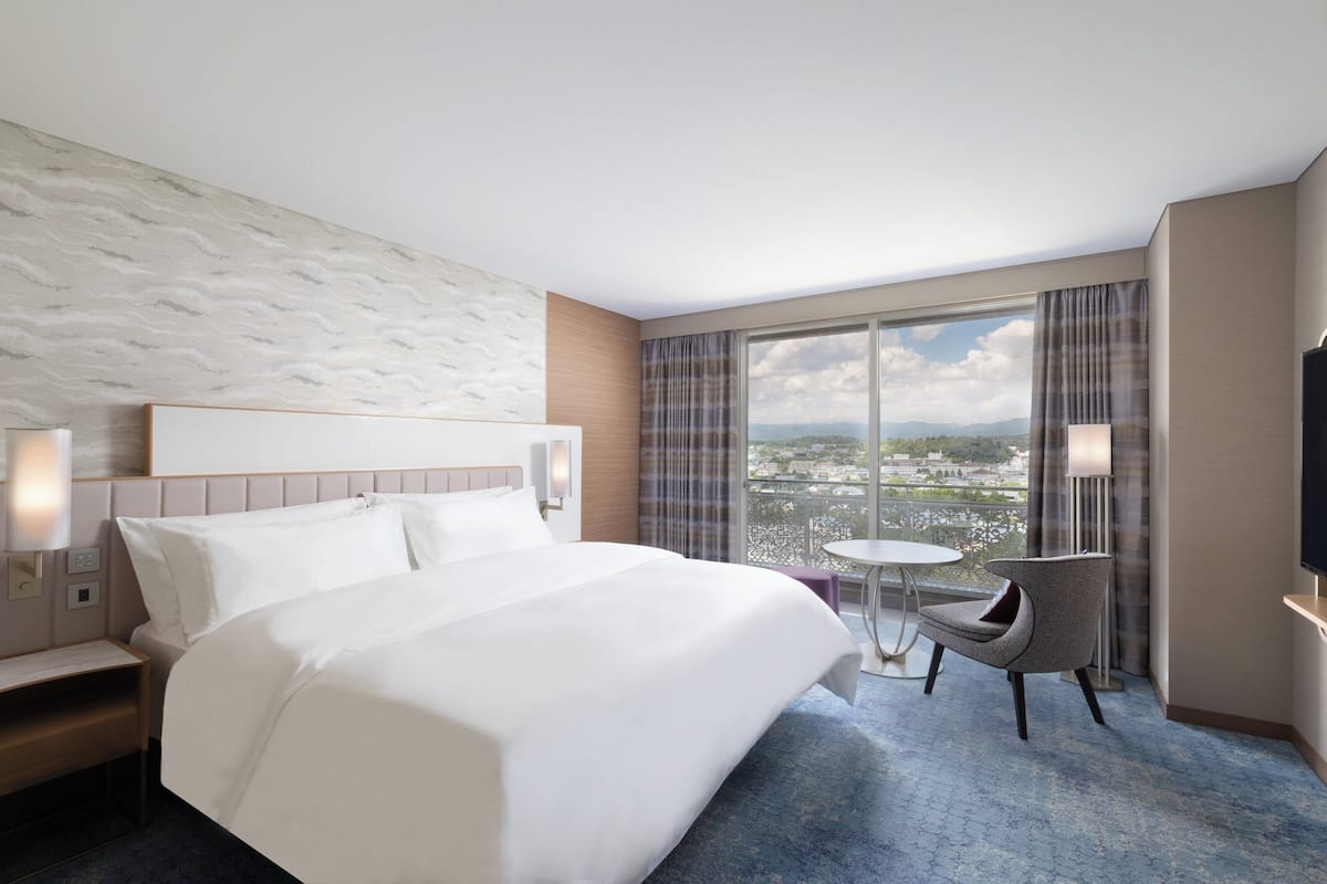 A hotel room with a large bed, a small round table, a chair, a TV, and a view of the city through large windows.