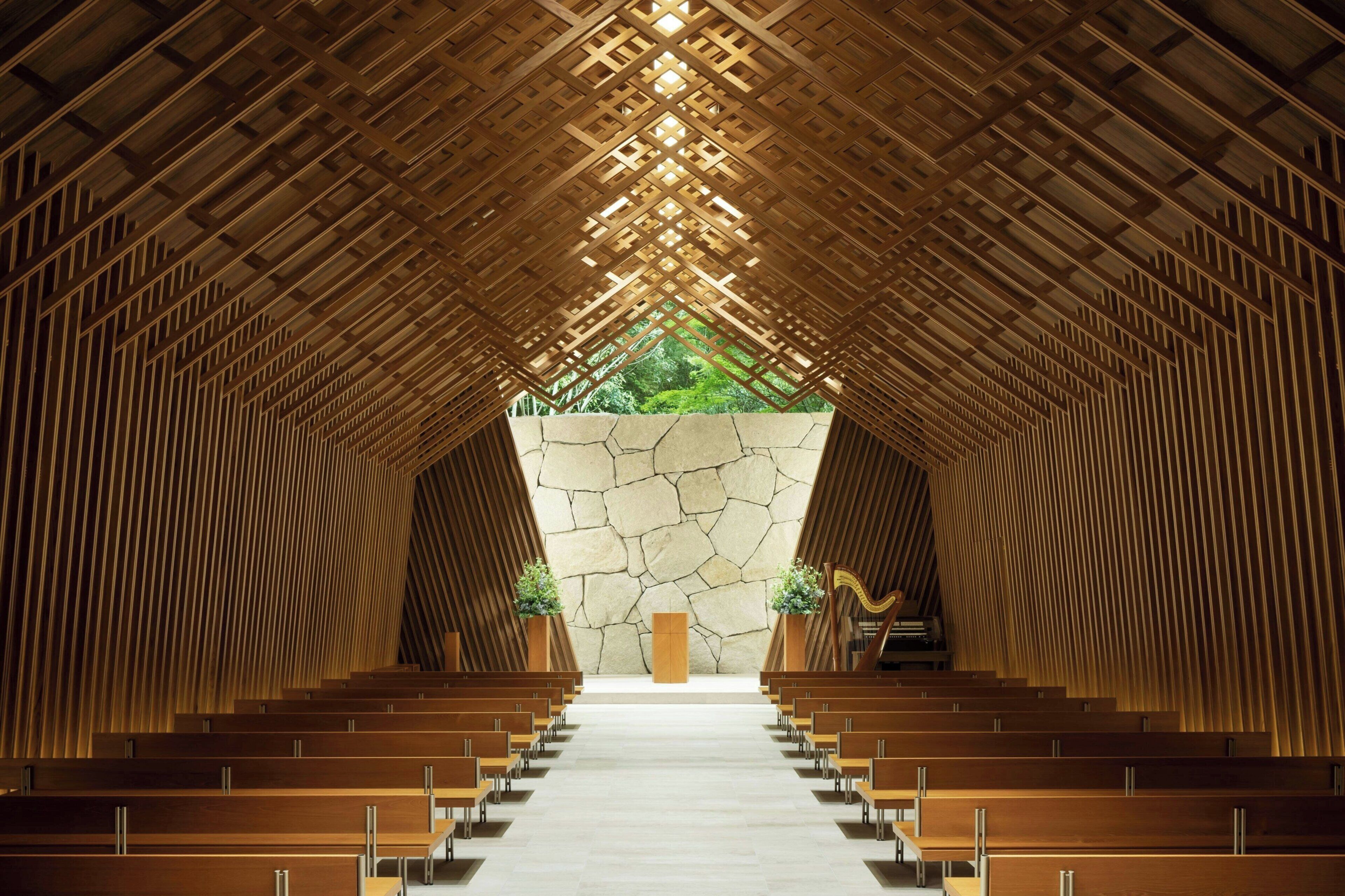 A church interior with wooden pews arranged in rows, a stone wall at the front, and a view of greenery outside.